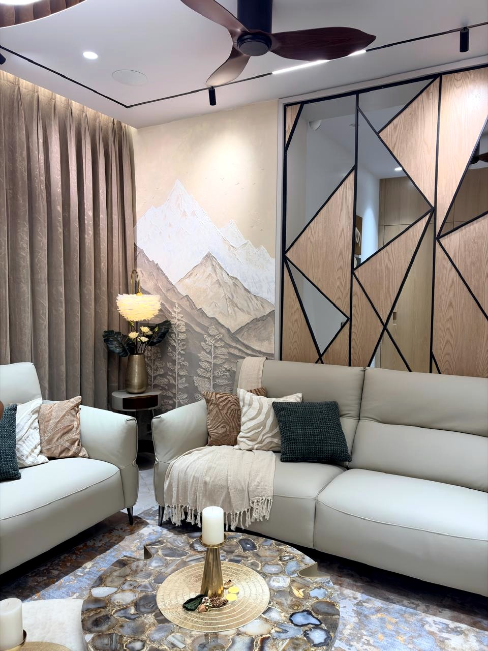 The living room of a Chennai apartment — a textured mountain mural behind cream sofas, a mirrored timber partition