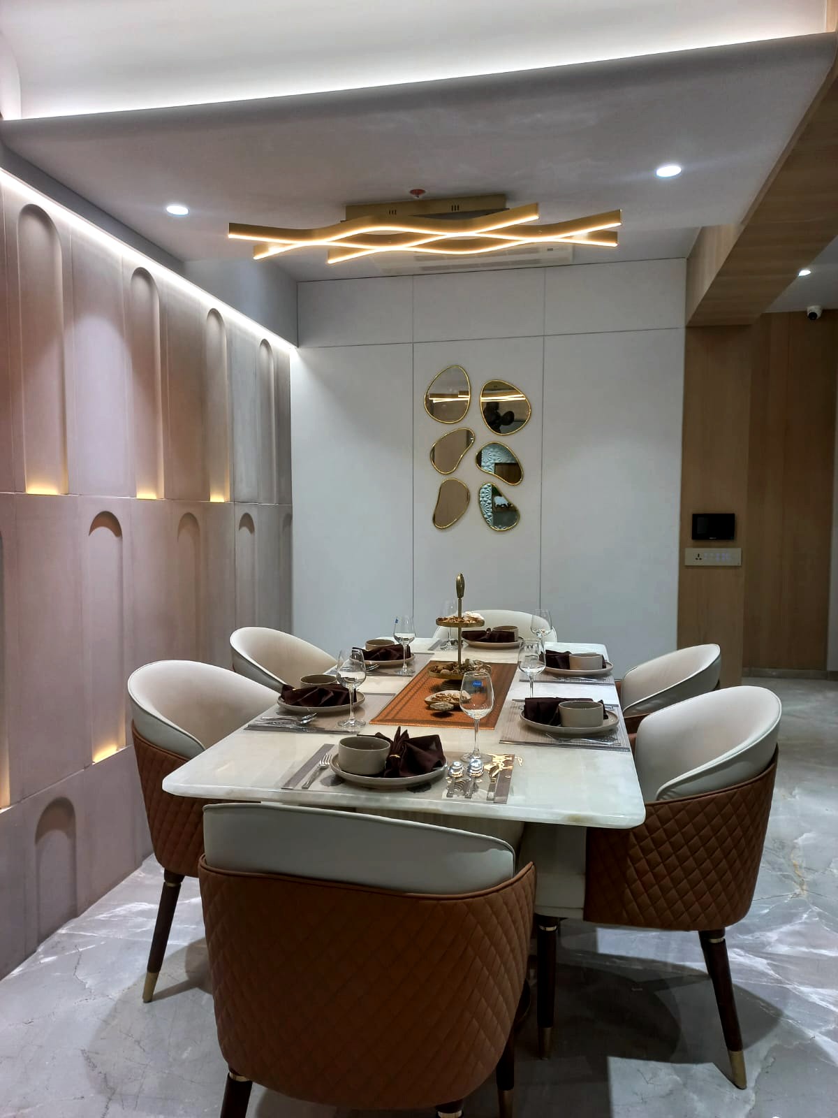 The dining room — arched fluted wall panels, a marble-topped table with tan quilted chairs, a cluster of organic gold-framed mirrors, a wave-form ceiling light