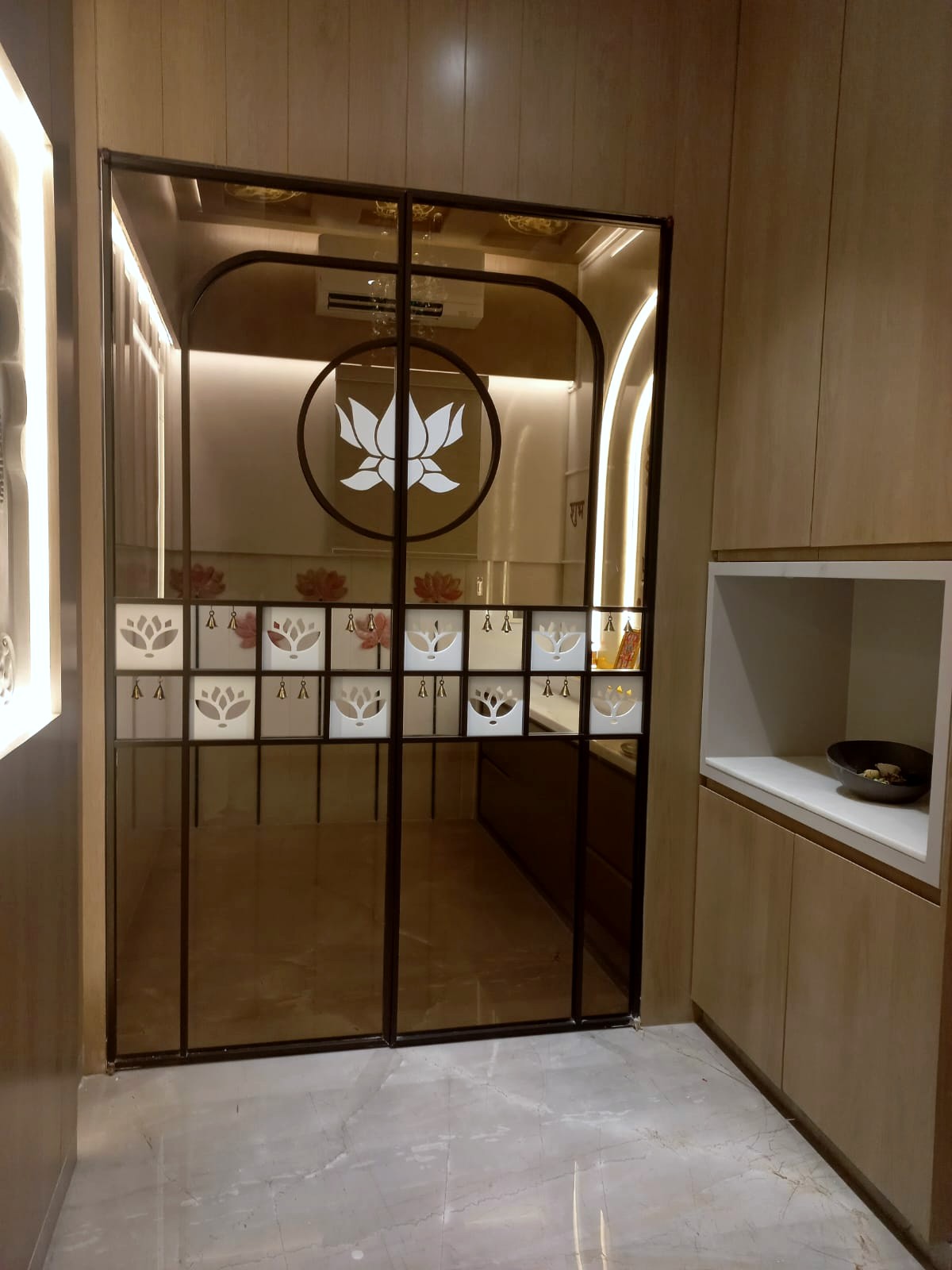 The mandir entrance — bronze-framed tinted glass sliding doors with an etched lotus medallion and a grid of lotus panels hung with small brass bells