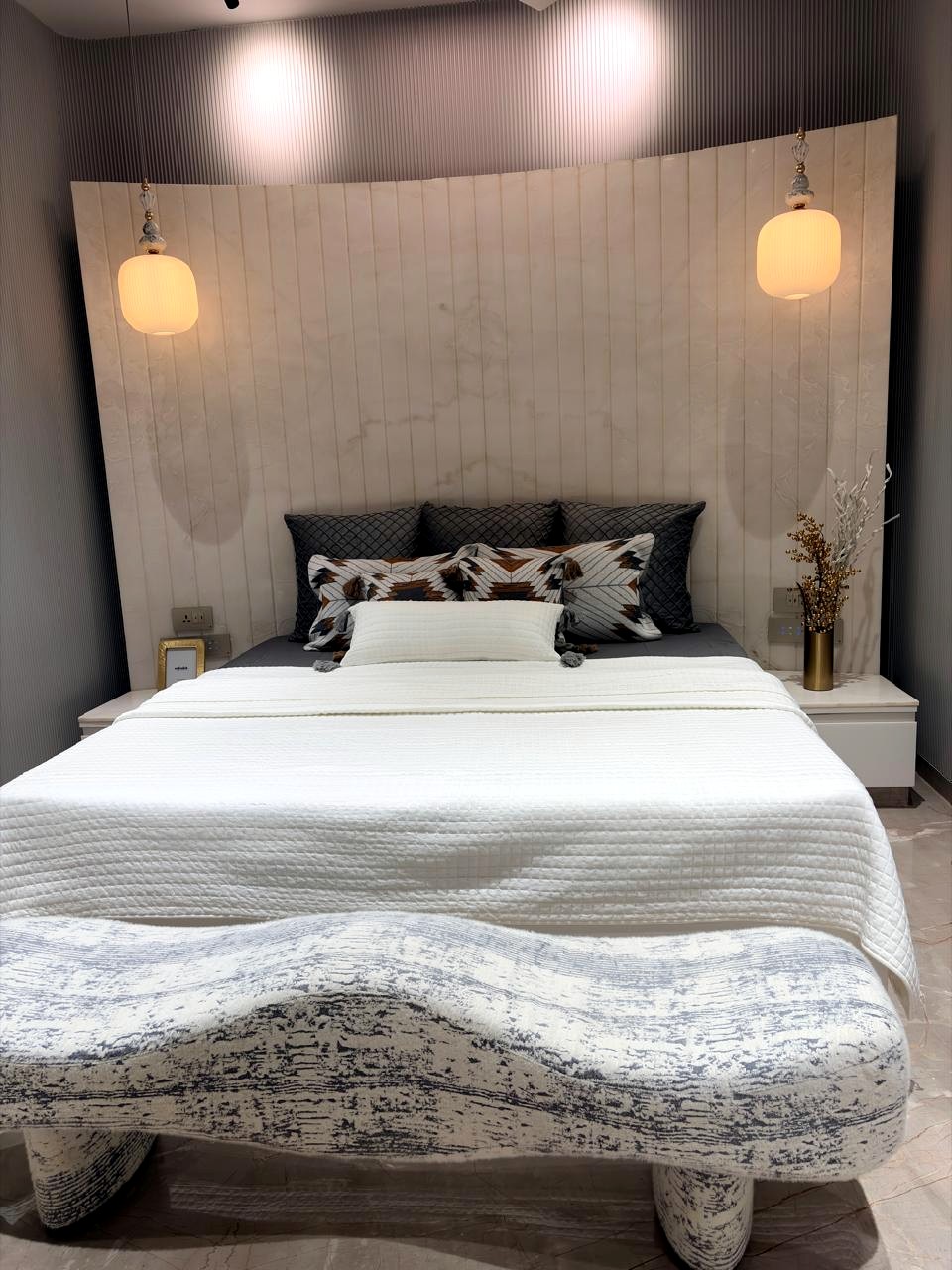 The parents' bedroom — a curved, fluted marble headboard wall, twin pendant lamps, grey and white bedding, soft beige palette