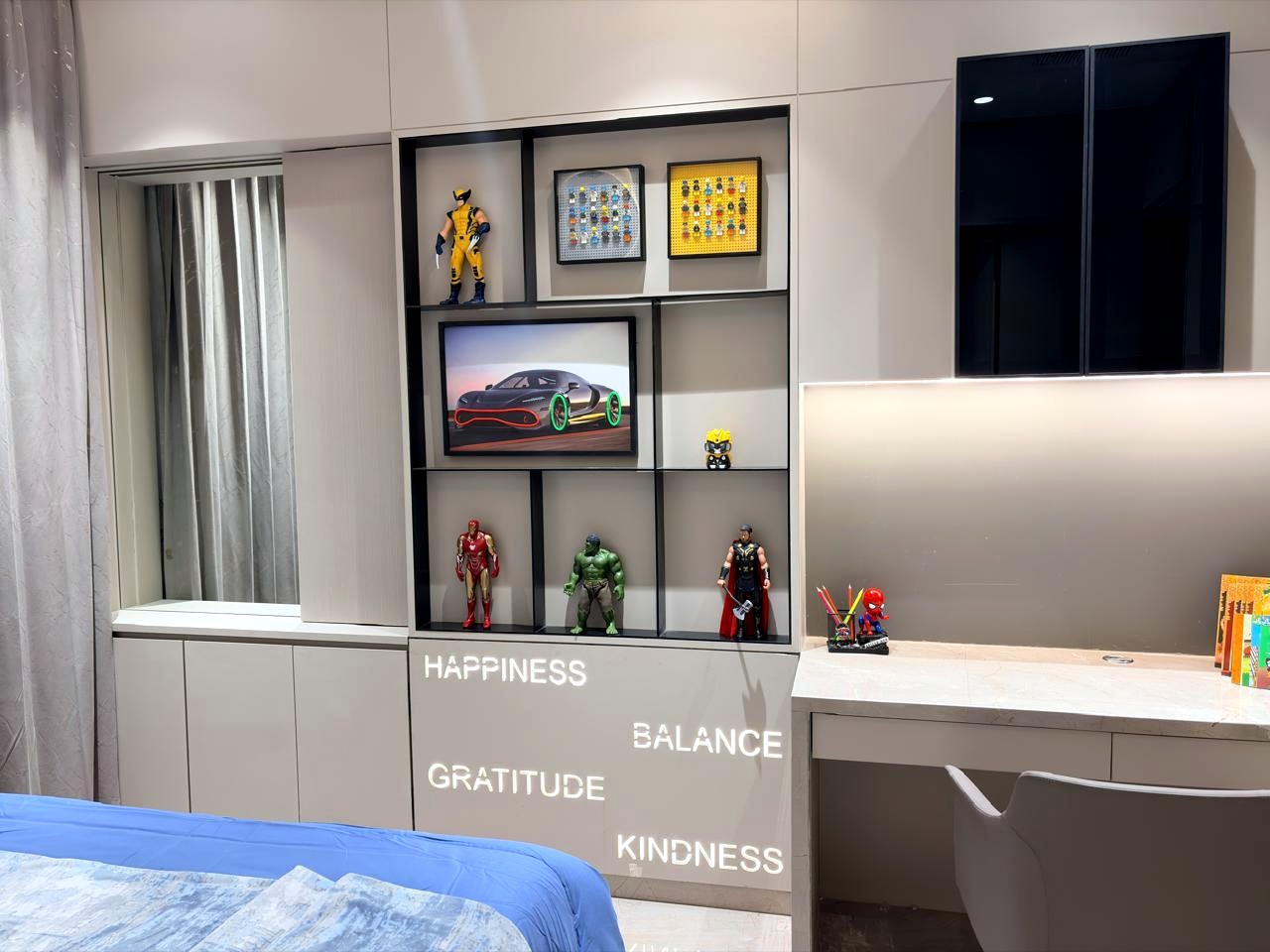 The son's display and study wall — open niches with superhero figures and Lego art, a built-in desk, the words happiness, balance, gratitude, kindness lit into the cabinetry