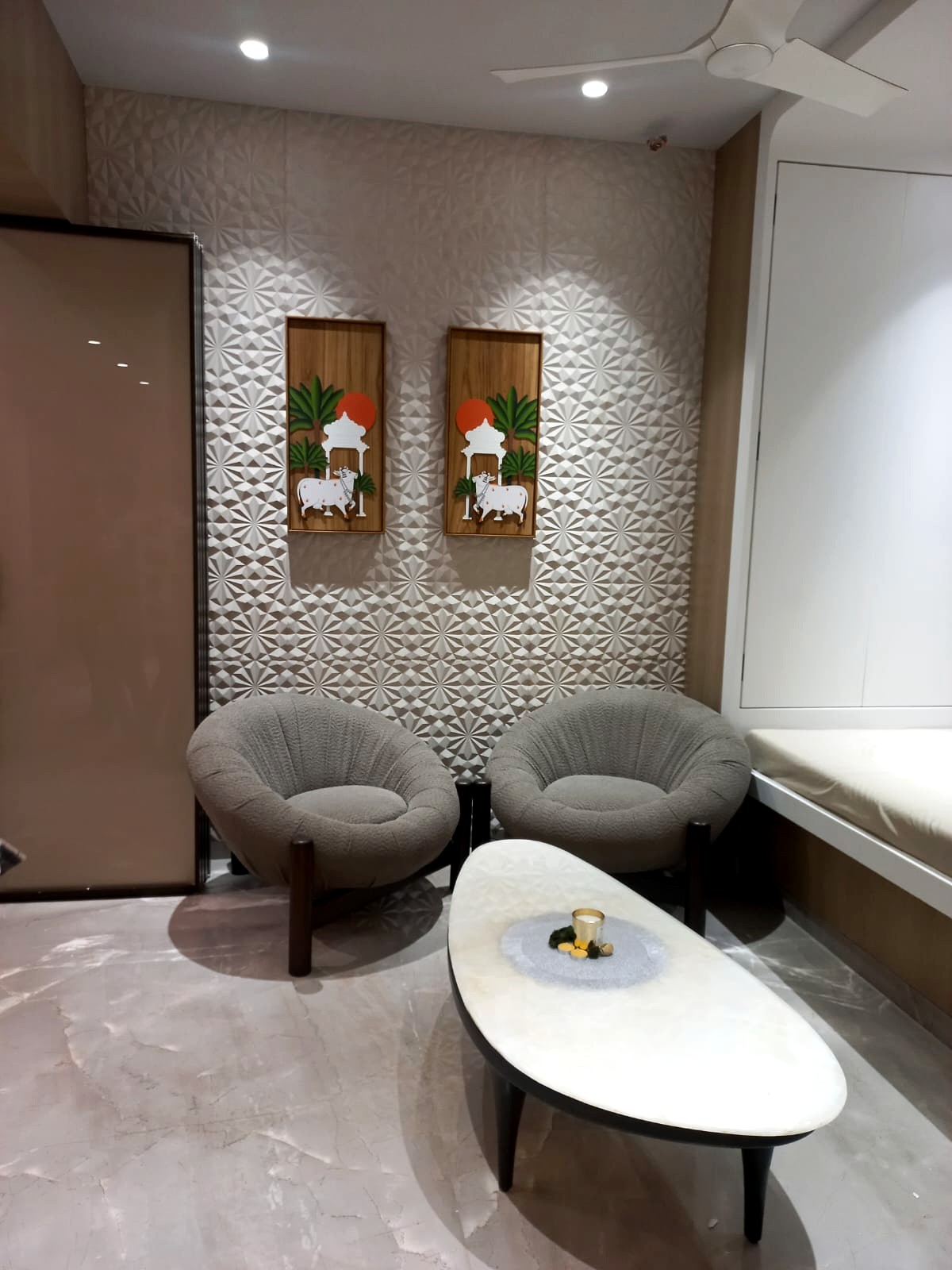 The family room — two grey tub chairs and a marble coffee table against a textured silver-grey panelled wall hung with two framed Pichwai cow artworks
