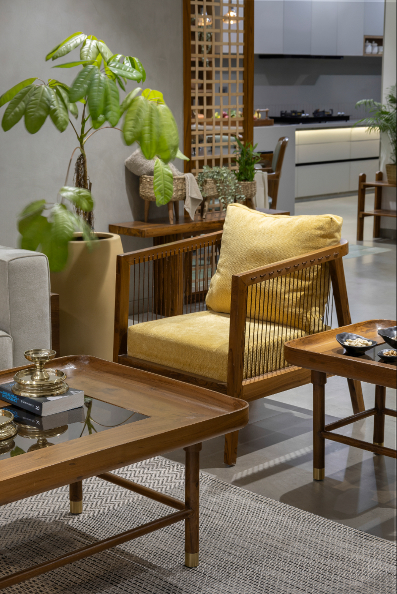 Detail of the living room — yellow cane armchair, teak side table, glass-topped coffee table, plants, kitchen visible through lattice