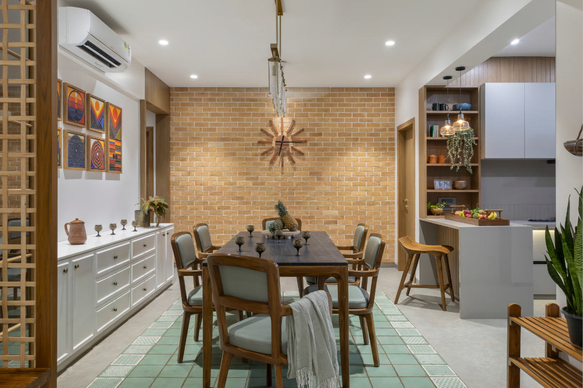 Wide view of the dining room — the exposed brick end wall with sunburst clock, dining table, kitchen and shelving visible beyond, lattice screen to the left