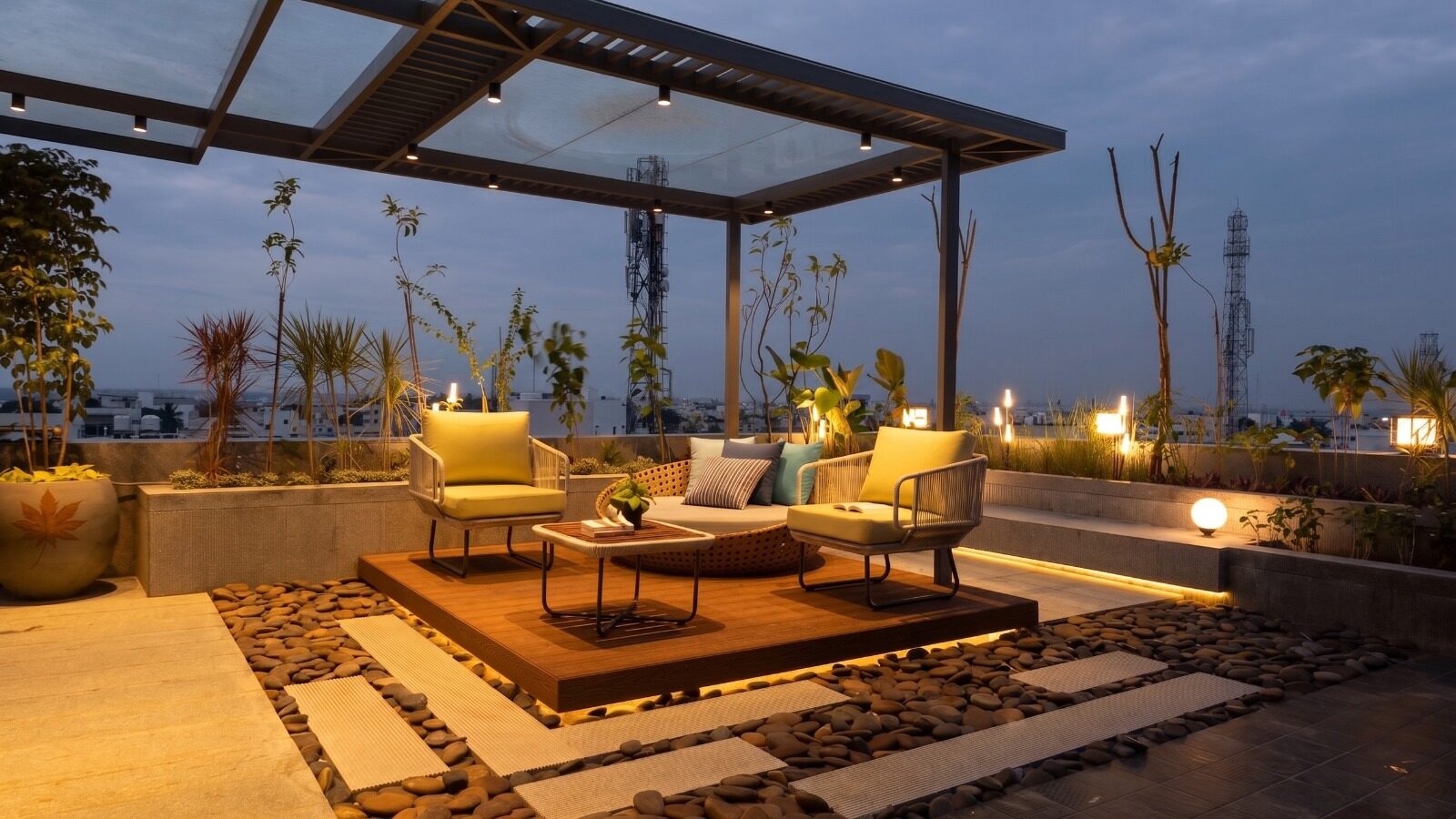 A pergola lounge on the terrace at twilight — woven chairs with yellow cushions, planters, mood lighting against an evening sky