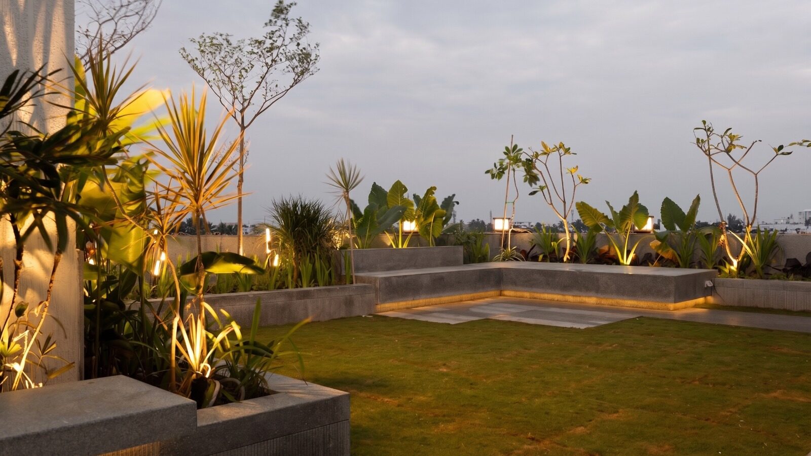 The terrace garden at dusk — a long cove-lit bench in the corner, planted beds with palms and frangipani against a blue evening sky