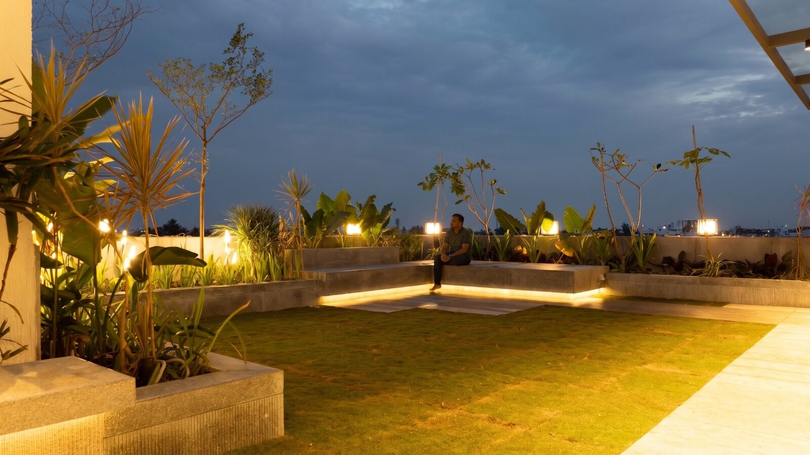 A wider view of the same terrace at dusk, one of the family members seated on the cove-lit bench, looking out across the city